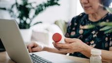 A close up of a woman holding a prescription bottle while using a laptop