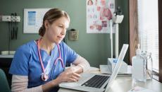 Healthcare provider wearing a stethoscope around their neck and scrubs sitting at a desk and looking at a computer