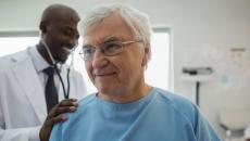 Healthcare provider in a lab coat standing behind a patient in a blue hospital gown checking their vitals