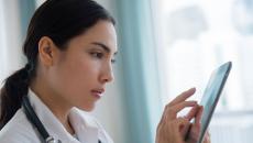 Healthcare provider wearing a lab coat with a stethoscope around their neck and looking at a tablet