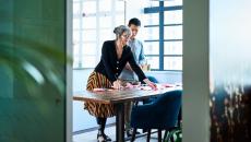 Two people standing in a room looking at something on a desk while standing above it 