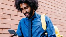 Person walking beside a brick wall wearing a blue shirt and yellow backpack while looking at a phone