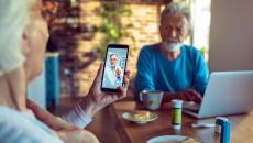 Two people sitting at a table with a computer on it and one of them is holding a phone