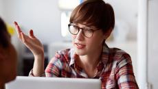 Person wearing glasses and a plaid shirt sitting at a computer with their hand up as though talking to someone on the computer