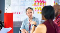 People sitting around a table in front of a whiteboard talking to each other 